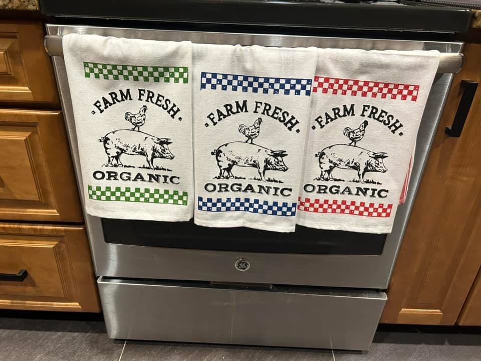 Three 'Farm Fresh Organic' towels hanging on a stove handle in a kitchen.