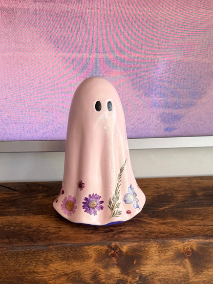 A pink ceramic ghost figurine with small black eyes and painted purple flowers, displayed on a rustic wooden mantelpiece in front of a pink patterned wall.