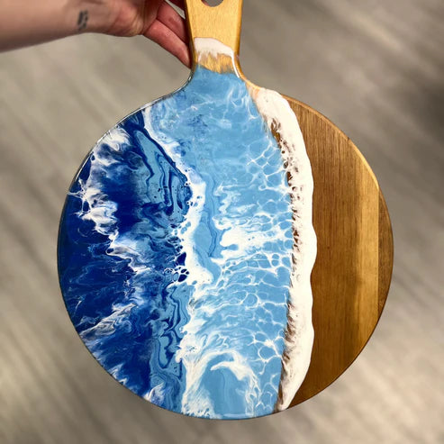 A round wooden serving board with a painted ocean wave design in blue, white, and navy, held by a hand over a neutral gray background.