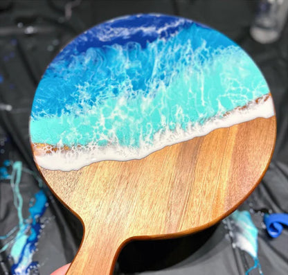 A close-up of a paddle-shaped wooden object, likely a spoon or small tool, featuring an ocean wave design in blue, turquoise, and white resin on the top surface.