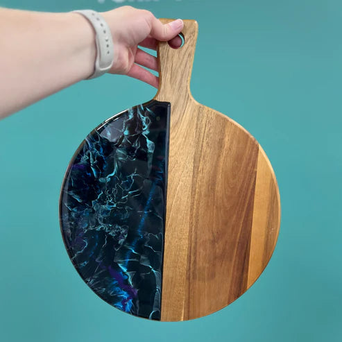 A hand holding a round wooden serving platter with a split design: one half is natural wood grain, and the other half features a glossy black and blue marbled pattern. The platter has a handle with a hanging hole and is set against a solid teal background.