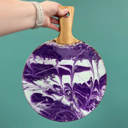 A hand holding a round serving platter with a wooden handle, featuring a purple and white marbled pattern with silver glitter accents, set against a solid teal background.
