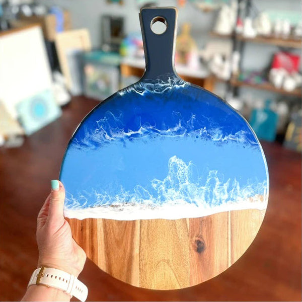 A hand holds a round wooden serving board with a dark blue handle. The board is decorated with a glossy resin design depicting blue ocean waves with white foam crashing onto a light wood surface.