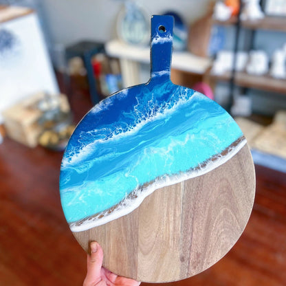 A round wooden serving board with a handle, featuring a resin ocean wave design in shades of blue, turquoise, and white, held by a hand in a home setting.