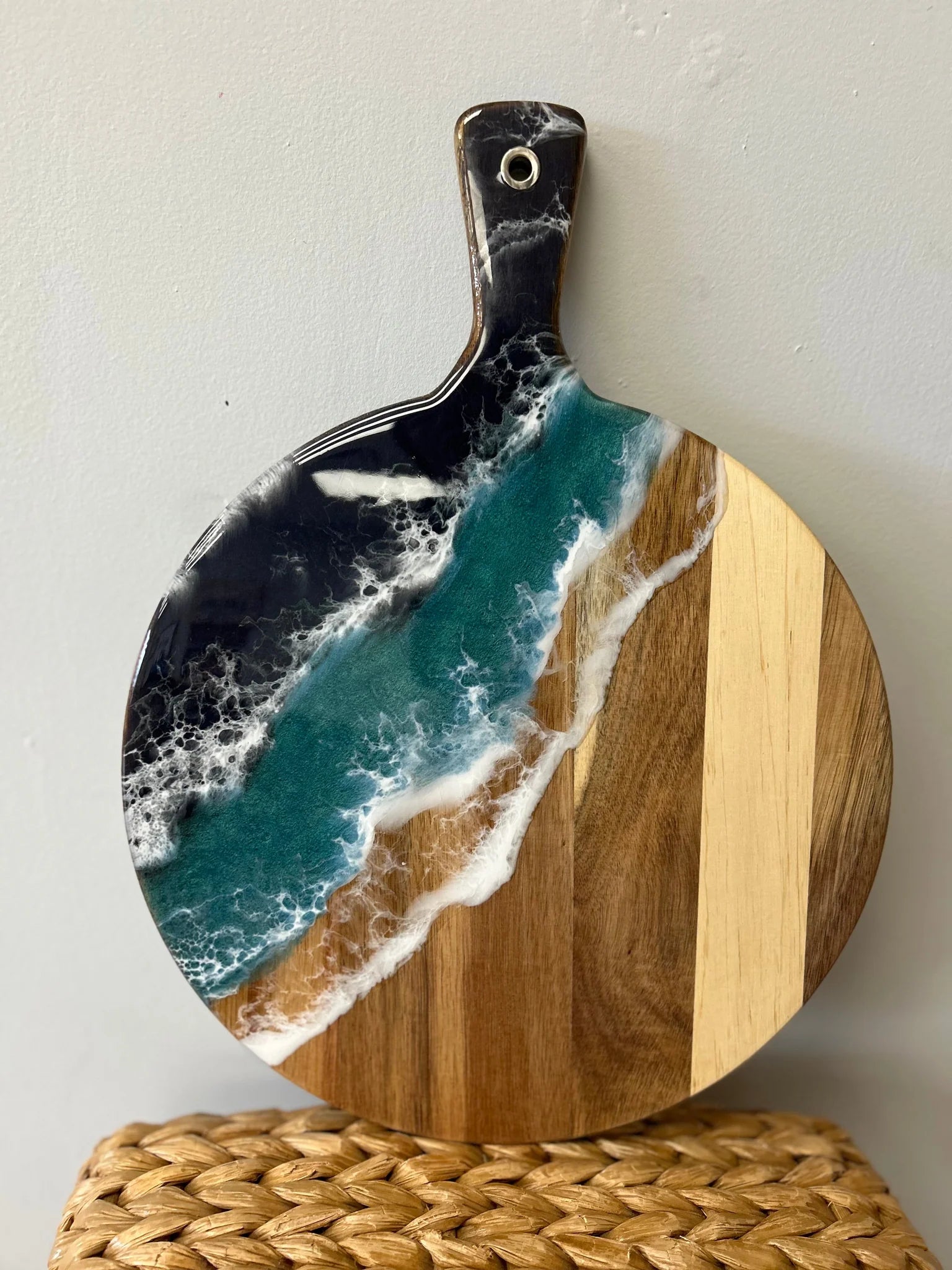 Round wooden serving board with a handle, featuring a printed or embedded ocean wave design in teal, white foam, and dark navy, with natural wood grain panels on the right side, displayed on a woven straw surface against a light wall.