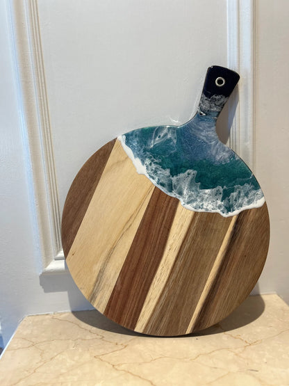 A round wooden cutting board with alternating light and dark wood stripes, featuring a wave-like resin inlay in teal, white, and blue around the handle, which is black with a white circular detail.