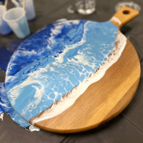 A round wooden serving board with a handle, featuring a resin art design of blue ocean waves and white foam on one half, placed on a dark tabletop.