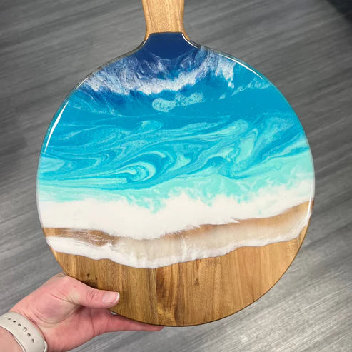 A hand holds a paddle-shaped serving board featuring a resin ocean wave design with blue and turquoise water, white foam, and a natural wood base and handle, set against a gray wood floor background.
