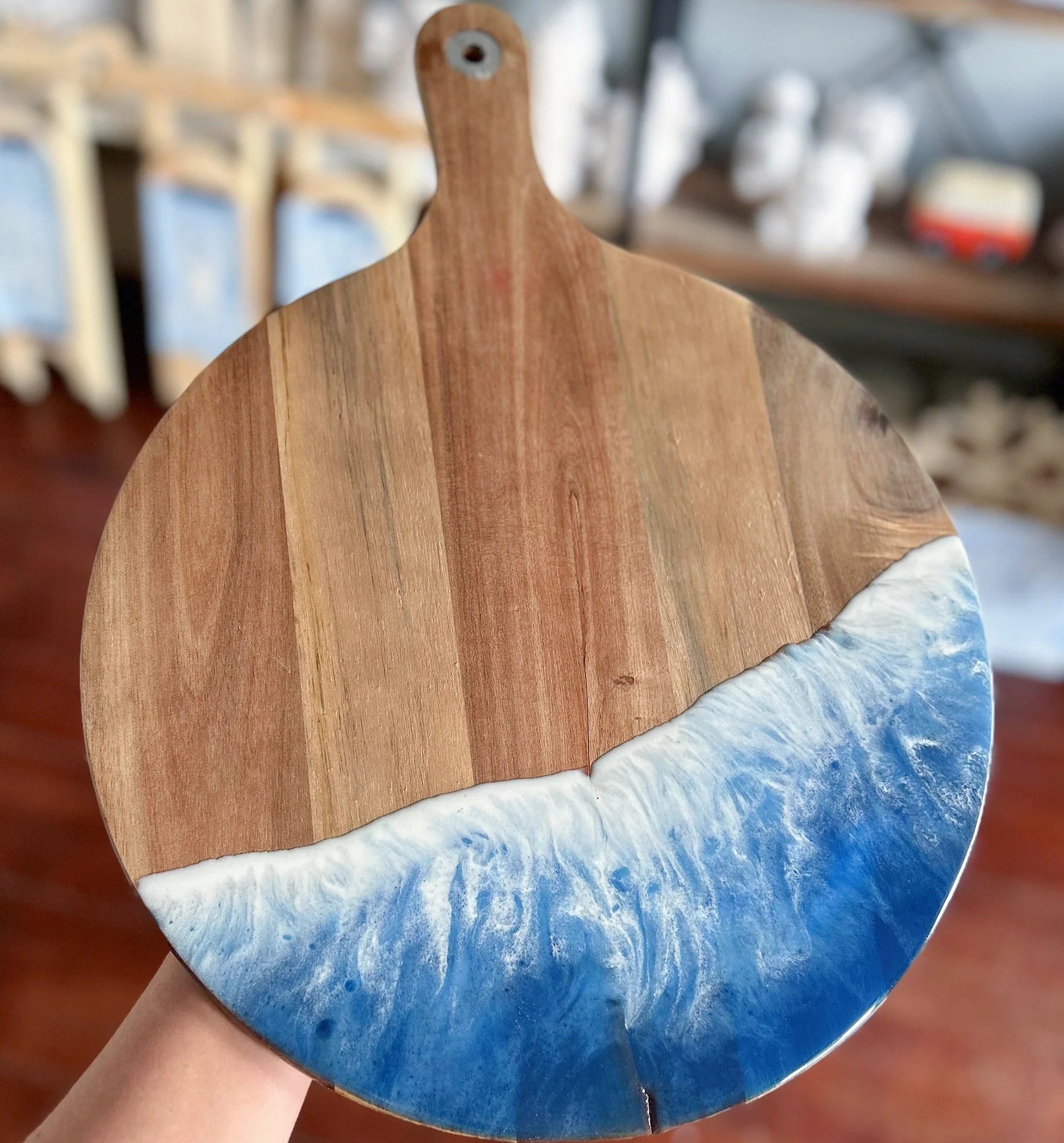A round wooden serving board with a natural wood grain top half and a blue and white resin wave design on the bottom half, held by a hand against a blurred indoor background.