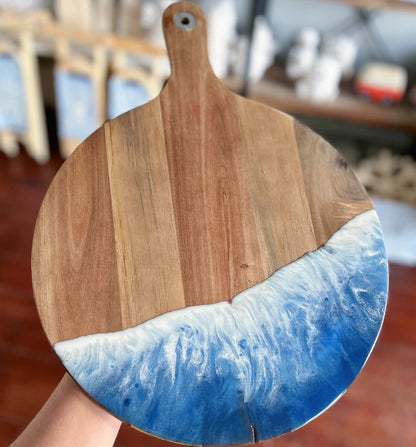 A round wooden serving board with a natural wood grain top half and a blue and white resin wave design on the bottom half, held by a hand against a blurred indoor background.