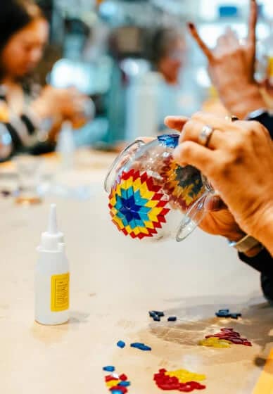 Person holding a colorful glass mug with a blurred background of people and objects.