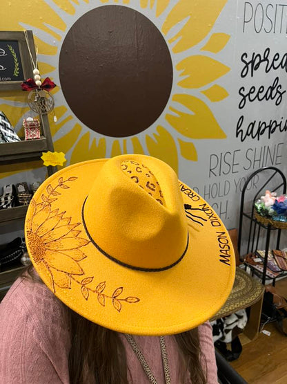Yellow hat with floral design worn by a person in front of a sunflower wall mural.