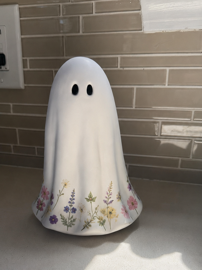 A white ghost-shaped ceramic container or candle holder with a floral pattern near the base, standing on a kitchen countertop against a tiled backsplash.