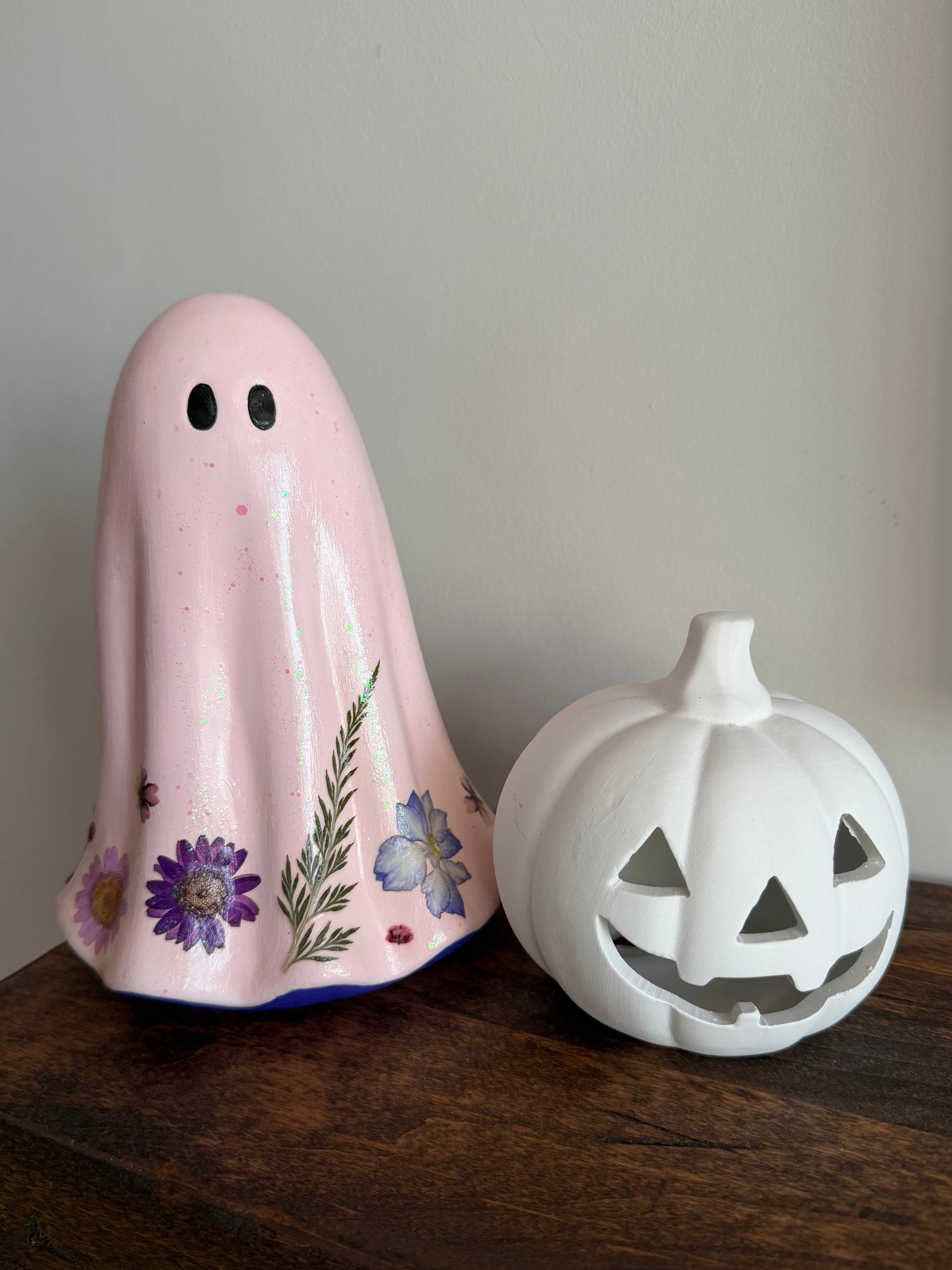A pink ghost-shaped ceramic candle holder decorated with purple and blue flowers, standing next to a white pumpkin-shaped candle holder on a dark wooden surface against a neutral wall.