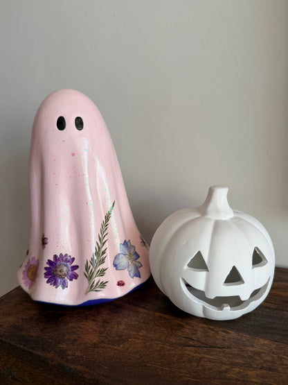 A pink ghost-shaped ceramic candle holder decorated with purple and blue flowers, standing next to a white pumpkin-shaped candle holder on a dark wooden surface against a neutral wall.