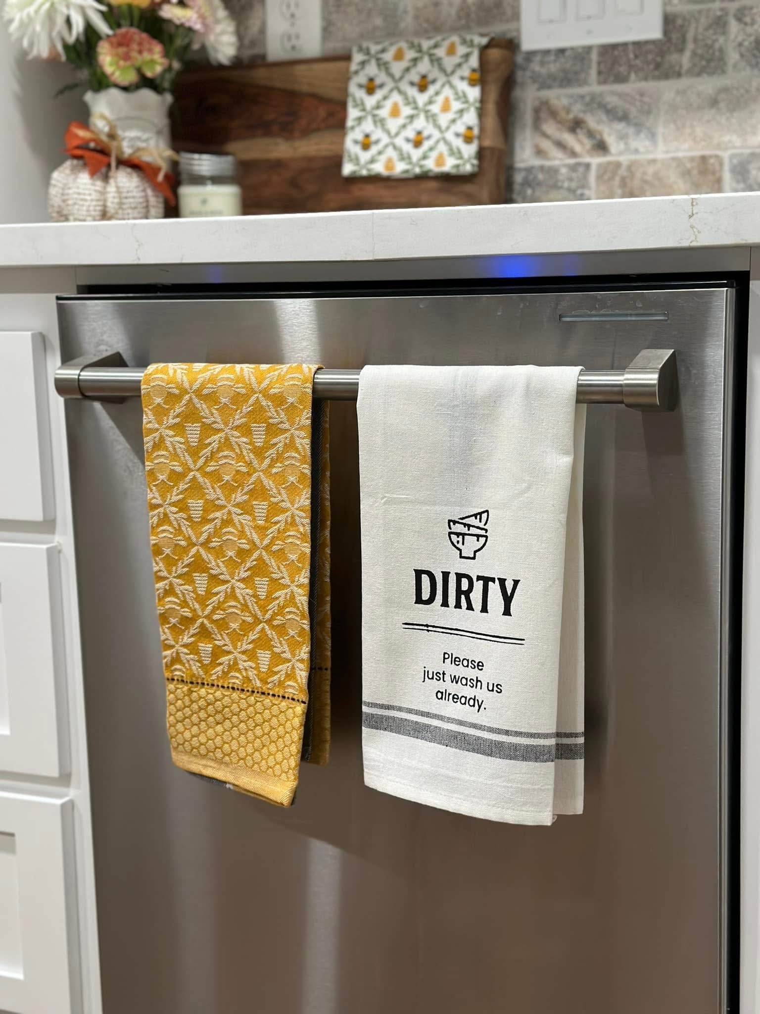 Two towels hanging on a kitchen towel rack, one yellow with a pattern and the other white with text.