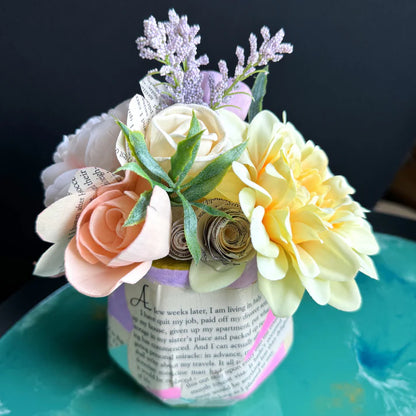 Decorative flower arrangement in a paper mache vase on a teal surface with a dark background