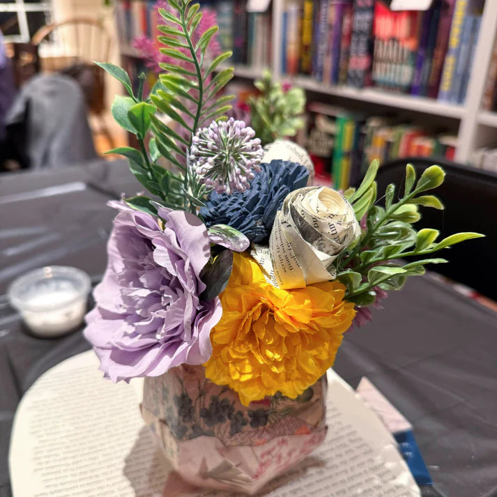 Bouquet of flowers with a yellow bloom and purple and green leaves on a table with books in the background.