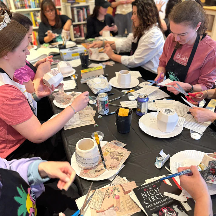 People sitting around a table with various items including cups, plates, and books in a casual setting.