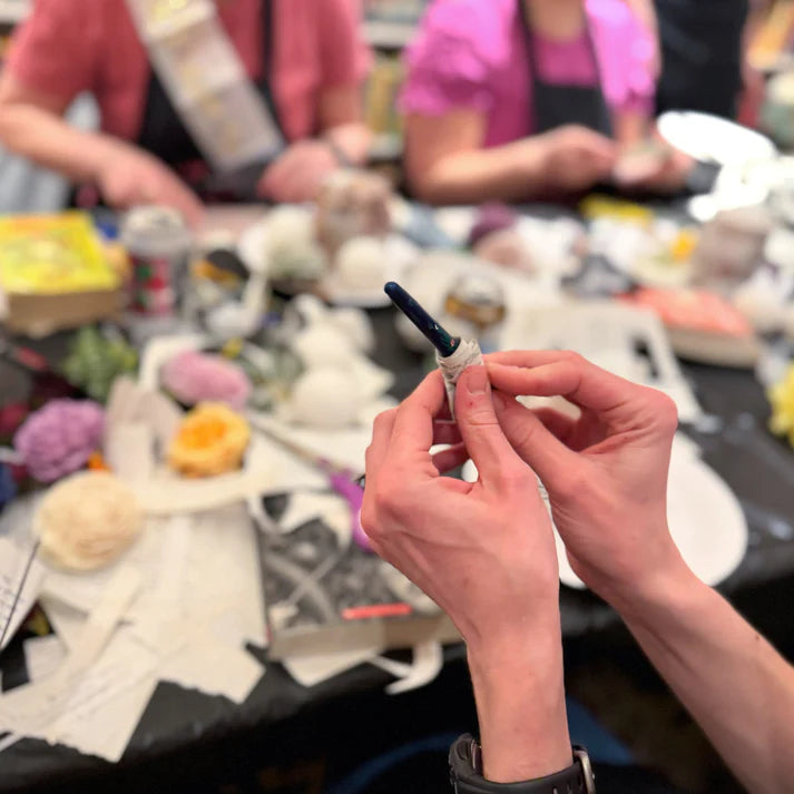Person holding a small object with a blurred background of people and items on a table.