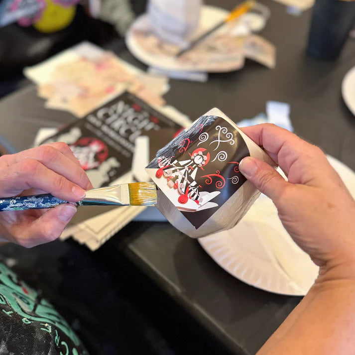 Person holding a small painting with a brush on a table with art supplies.