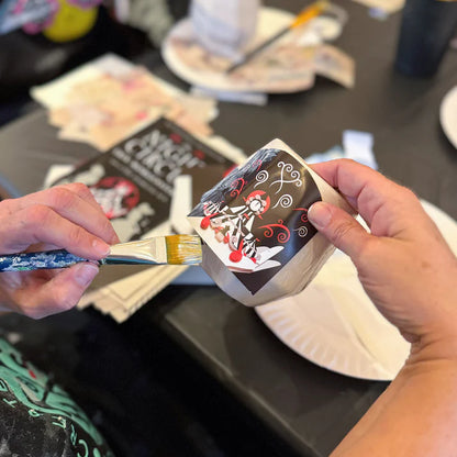 Person holding a small painting with a brush on a table with art supplies.