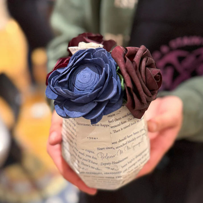 Hand holding a small vase with blue and burgundy flowers against a blurred background
