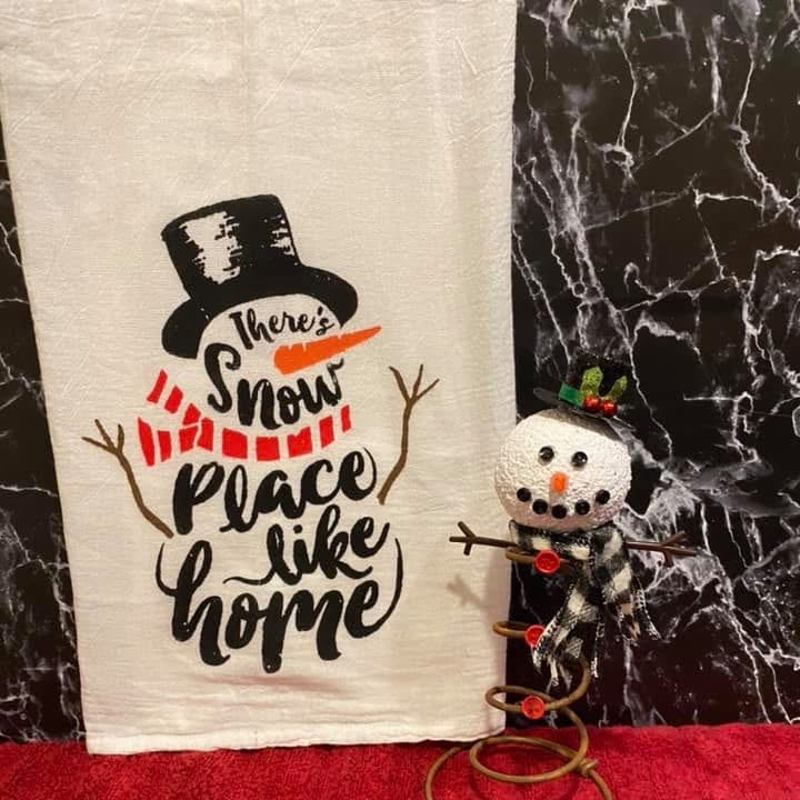 Decorative towel with snowman design and text, next to a small snowman figurine on a red surface.