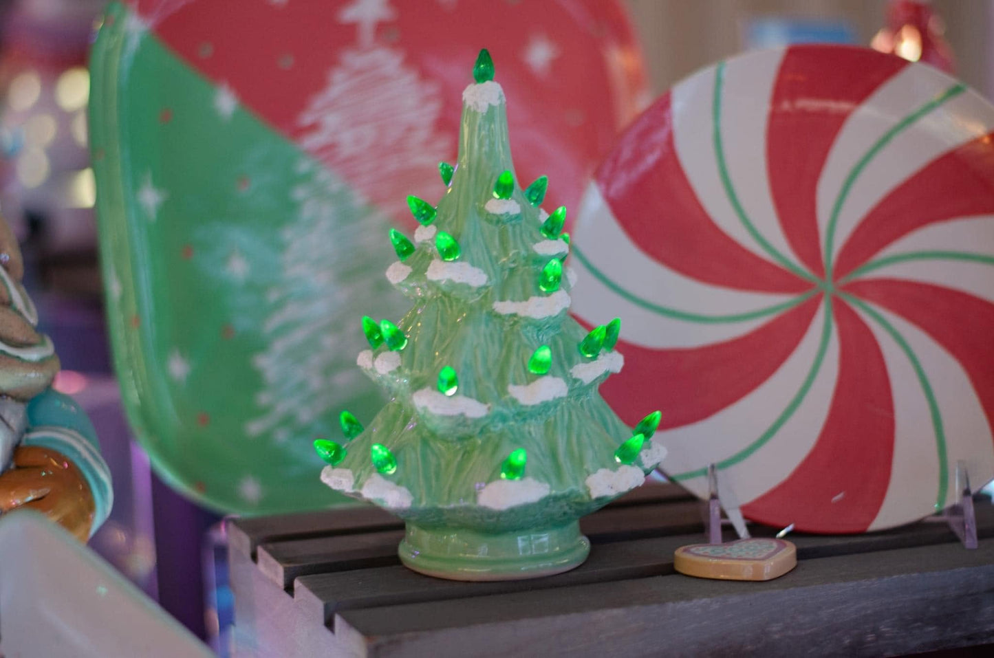 A small green ceramic Christmas tree ornament with glowing green lights and white snow-like accents, displayed on a dark wooden surface in a festive setting with candy cane decorations in the background.