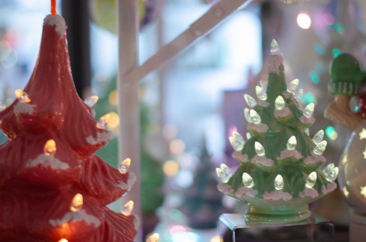 A close-up of a red Christmas tree-shaped ornament or candle with warm glowing lights, next to a green Christmas tree-shaped lighted ornament, in a softly blurred festive setting.