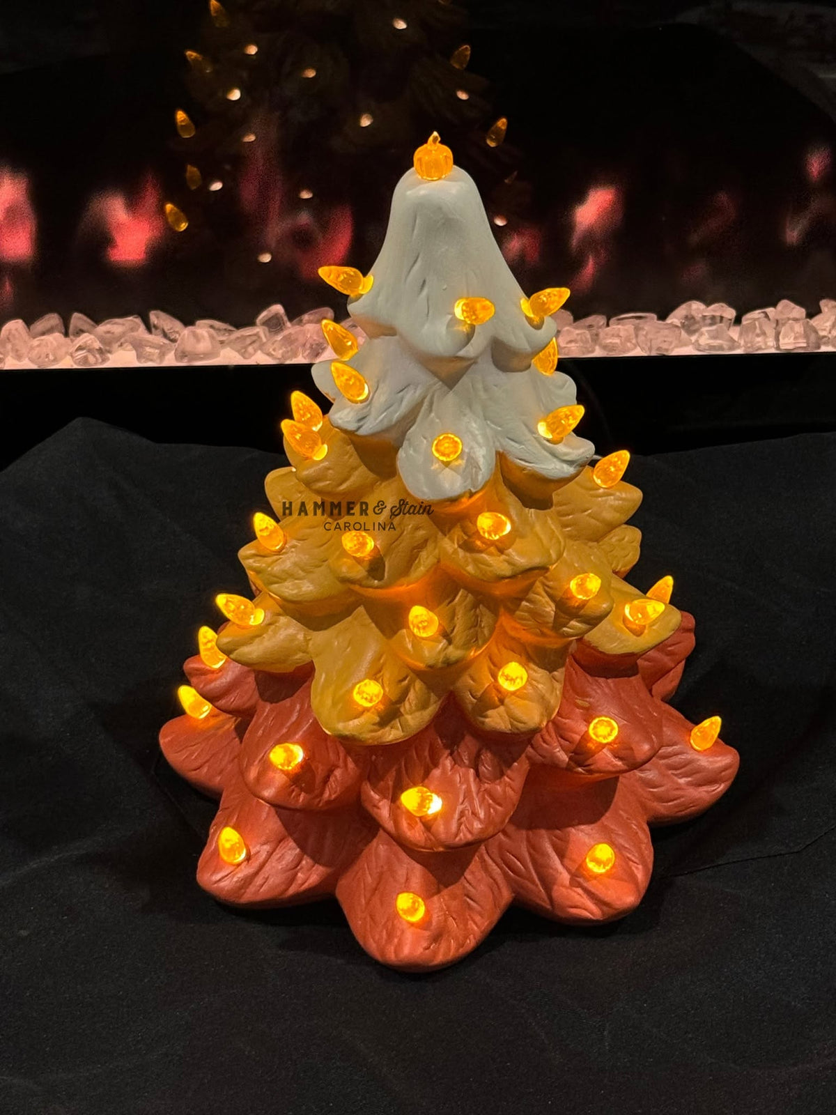 A decorative tiered tree sculpture with warm white LED lights glowing from within, set against a dark background with a blurred fireplace-like scene.