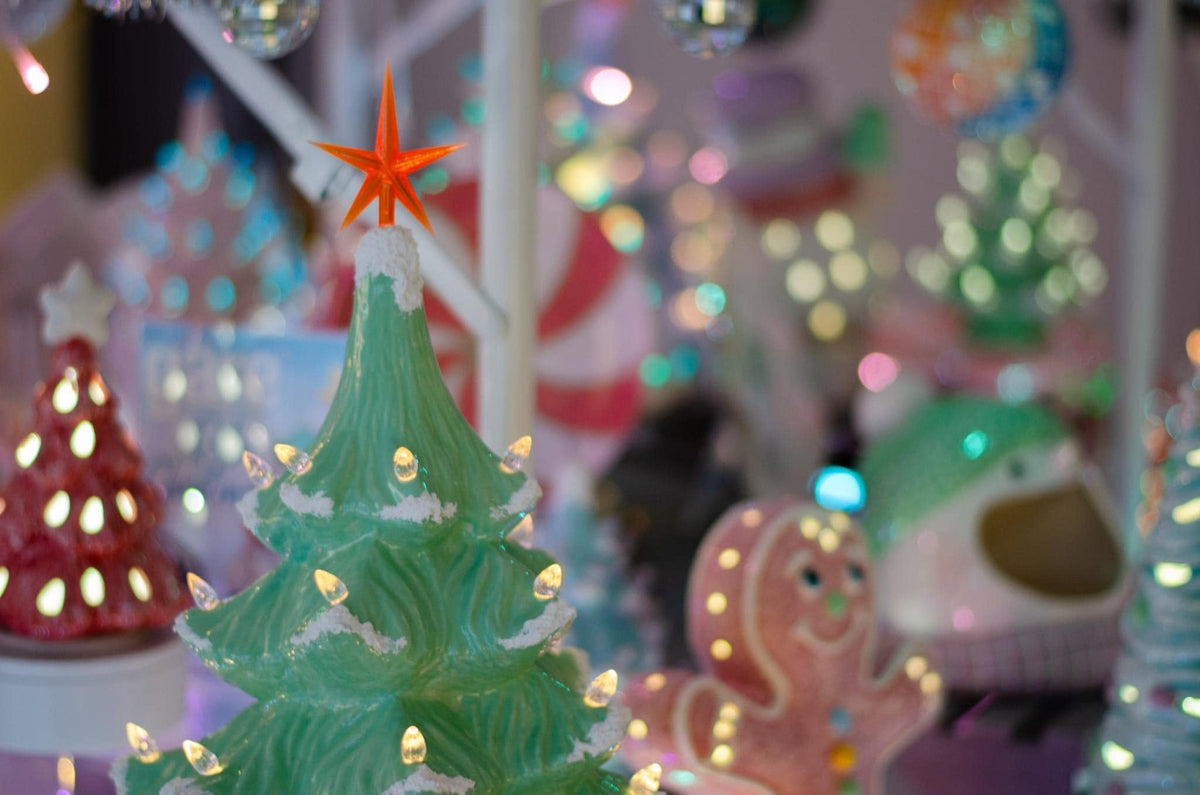 A close-up of a green Christmas tree ornament with a glowing warm white star and lights, surrounded by softly blurred festive decorations in the background.