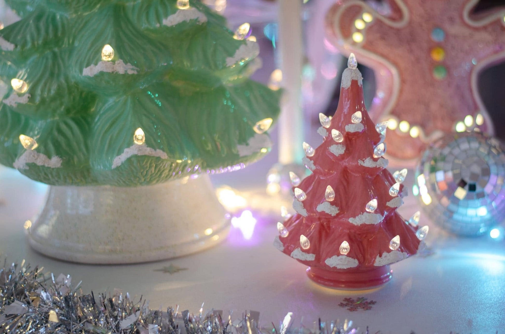 A vintage-style pink ceramic Christmas tree ornament with glowing white lights and silver snow accents, displayed next to a larger green tree on a tabletop with holiday decorations in the background.