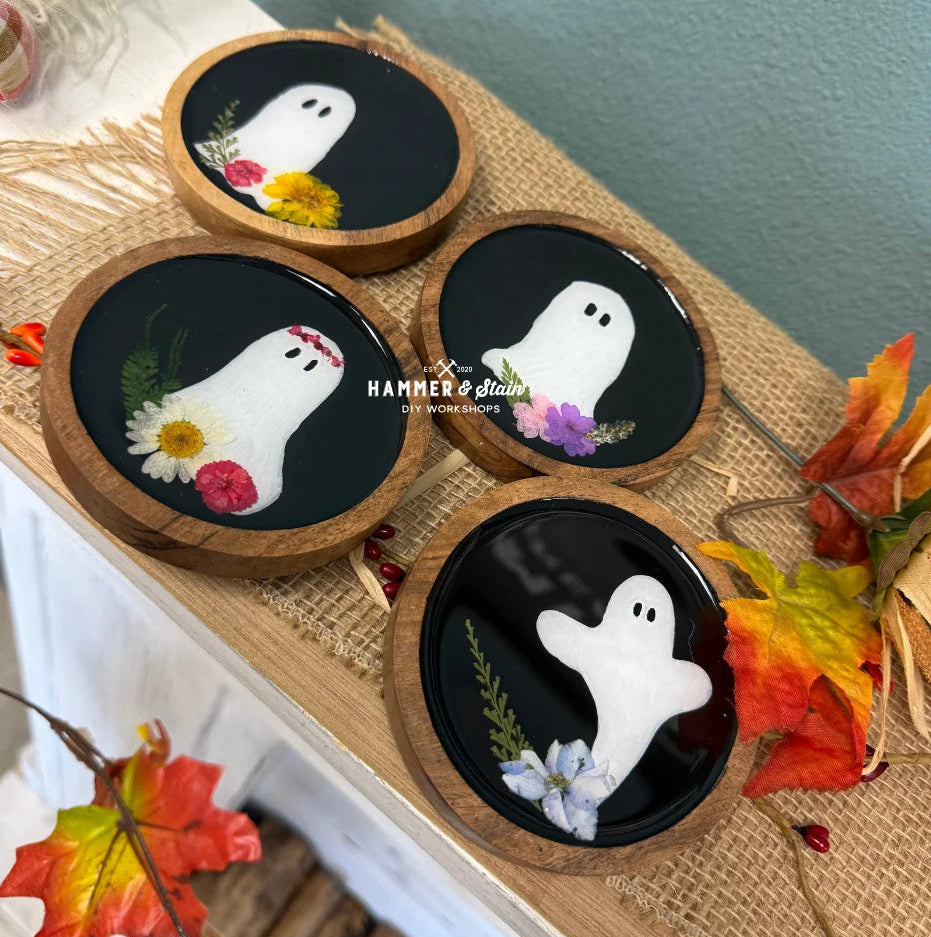 Three round wooden coasters with black centers featuring white ghost illustrations and small pressed flowers, arranged on a burlap runner with autumn leaves. A watermark reads Hammero Glazes.