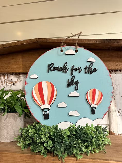 Decorative sign with hot air balloons and 'Reach for the sky' text on a wooden surface.