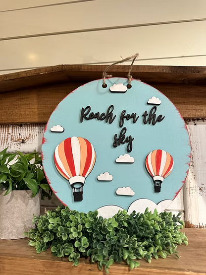 Decorative sign with hot air balloons and 'Reach for the sky' text on a wooden surface.