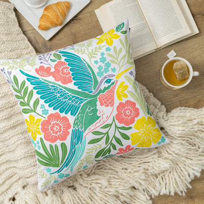 Decorative pillow with a colorful hummingbird and floral design on a wooden table with tea and pastries.