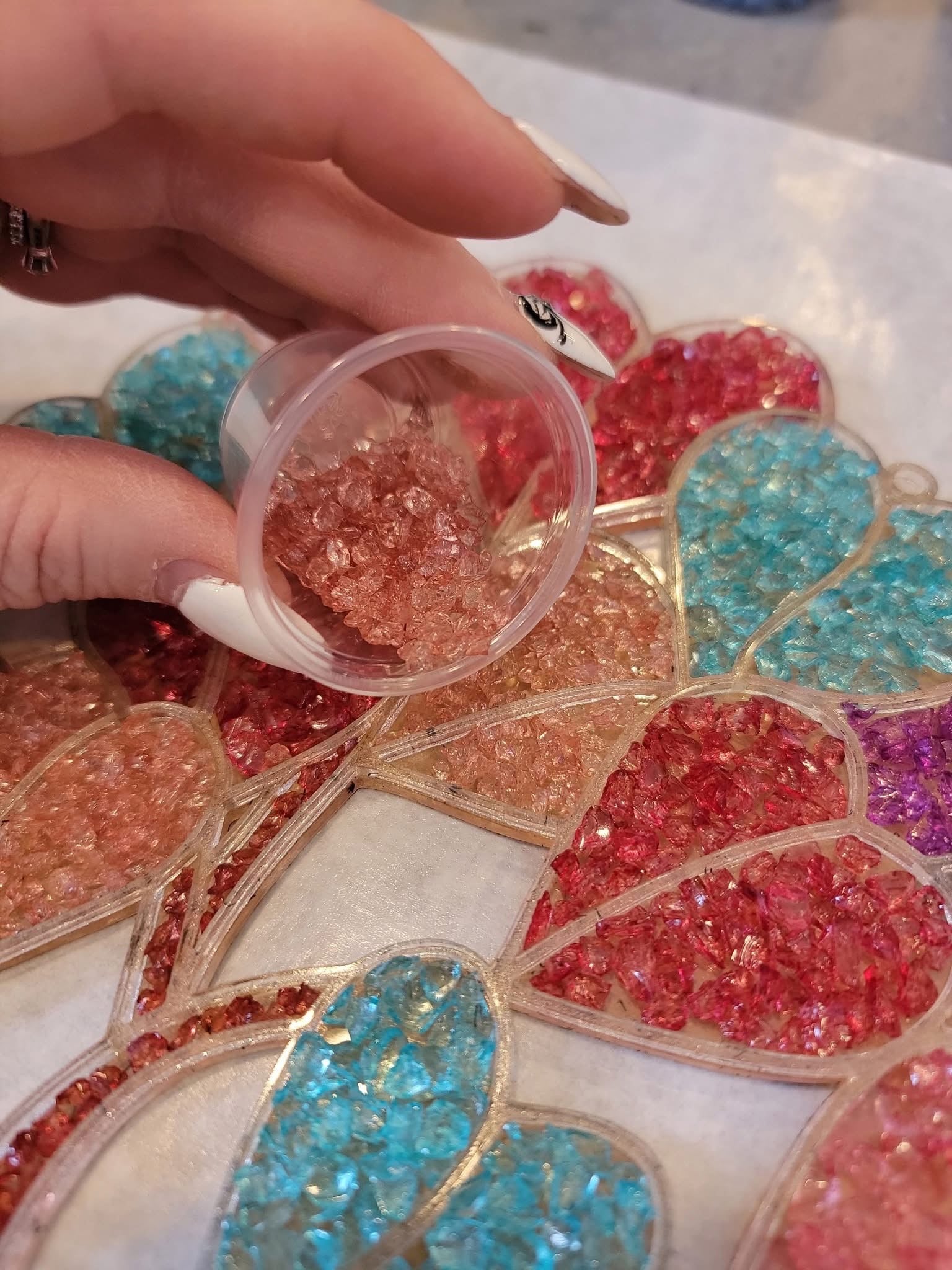 Hand holding a small container of crushed glass over a decorative surface with heart shapes filled with colorful crushed glass.