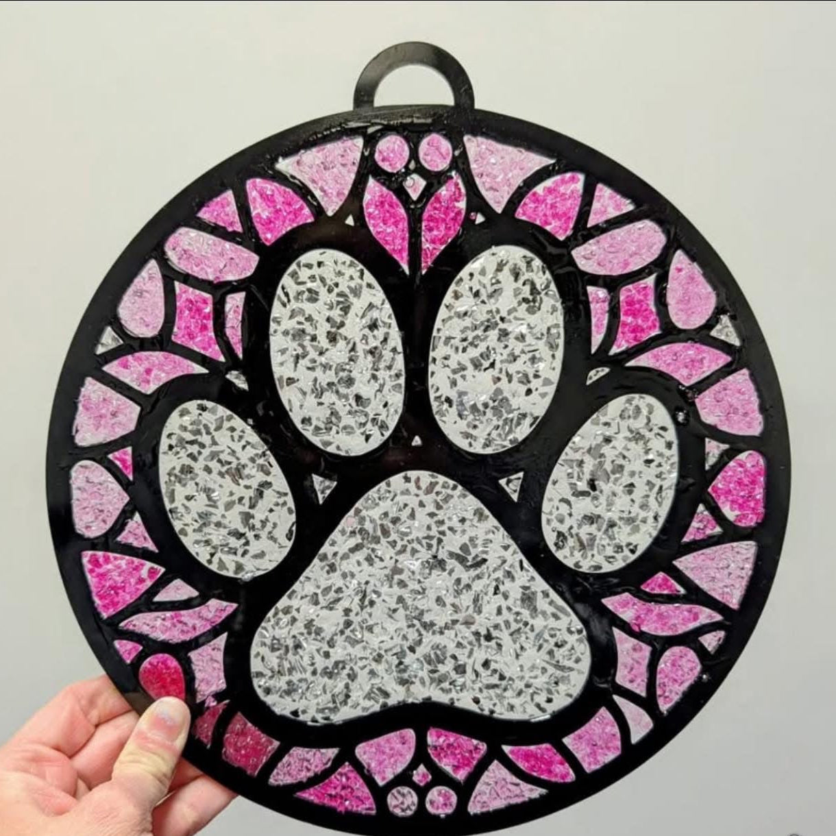 Decorative pet paw print with pink and black border held by a hand on a gray background