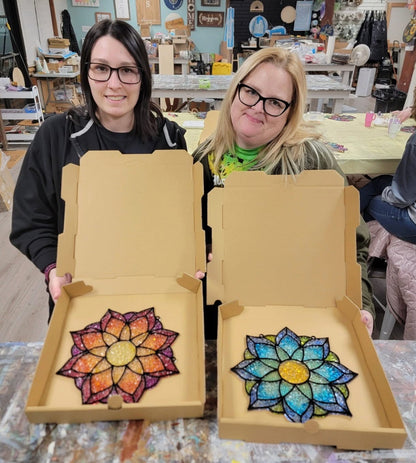 Two women holding open boxes with colorful mandala designs inside, in a workshop setting.