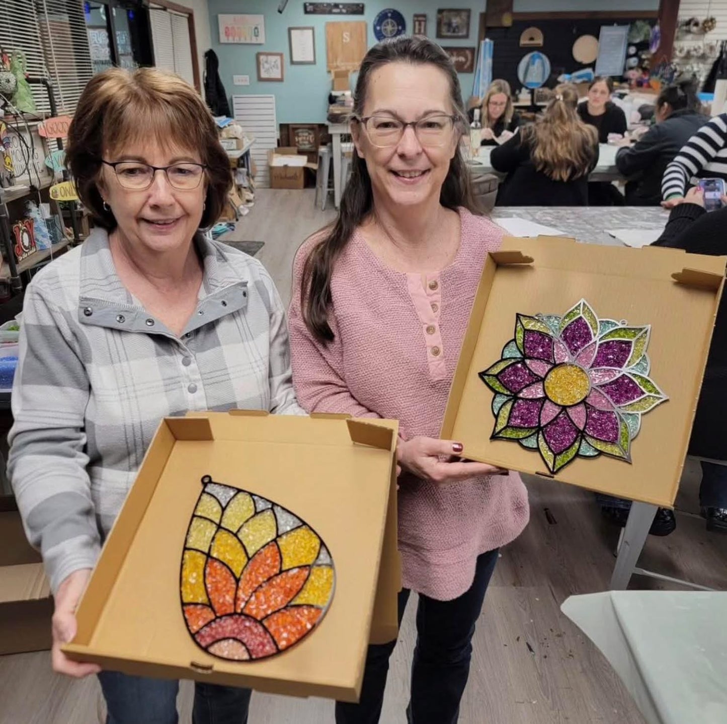 Two women holding colorful mandala and flower art pieces in a classroom setting.