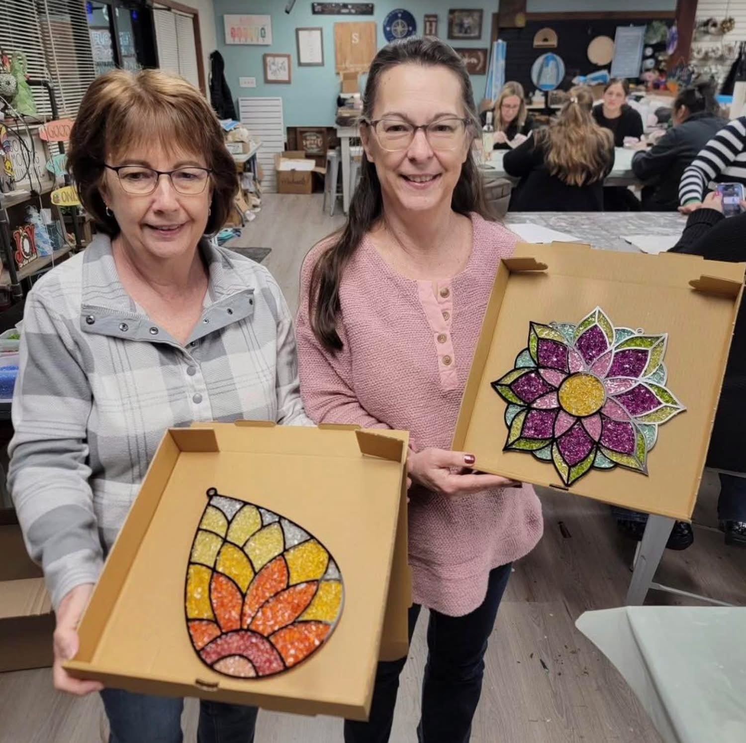 Two women holding colorful mandala and flower art pieces in a classroom setting.