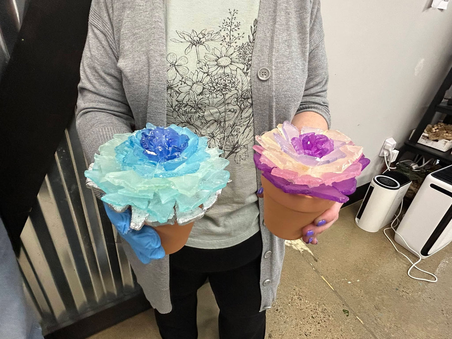 Person holding two colorful sea glass flower-shaped objects in pots, one blue and one pink, in an indoor setting.