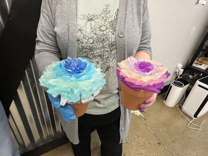 Person holding two colorful sea glass flower-shaped objects in pots, one blue and one pink, in an indoor setting.