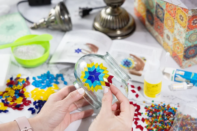 Person holding a glass jar with colorful star-shaped objects on a table with craft supplies.