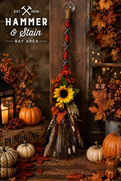 A decorative hanging broom with a red and black braided handle, a clear hanging loop, and a autumn-themed floral and straw raffia lower section, displayed in a styled fall setting with pumpkins, leaves, and warm string lights. 