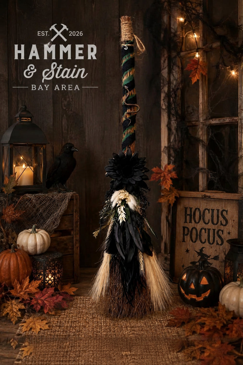 A Halloween decorative broom with black feathers, white feather accents, and a striped handle, styled in a rustic autumn scene with pumpkins, lanterns, and a wooden sign reading 'Hocus Pocus'.