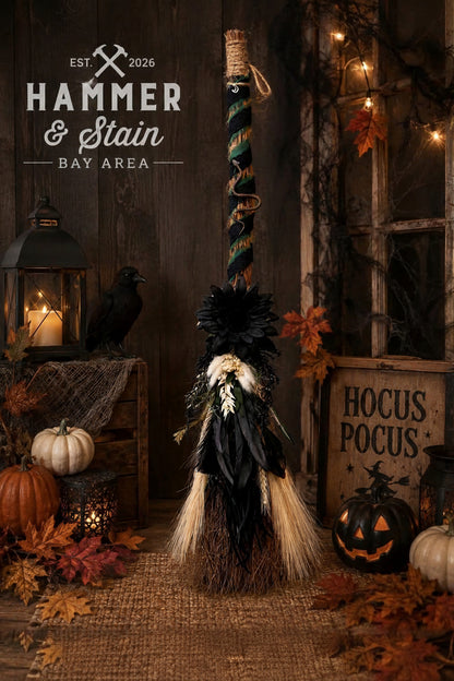 A Halloween decorative broom with black feathers, white feather accents, and a striped handle, styled in a rustic autumn scene with pumpkins, lanterns, and a wooden sign reading 'Hocus Pocus'.