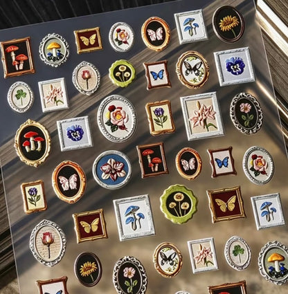 A sheet of small decorative enamel pins or charms featuring floral, butterfly, mushroom, and geometric designs in assorted colors, arranged on a dark reflective surface.