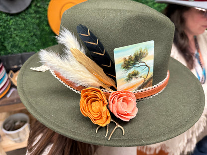 Green hat with feathers and flowers worn by a person, with a blurred background